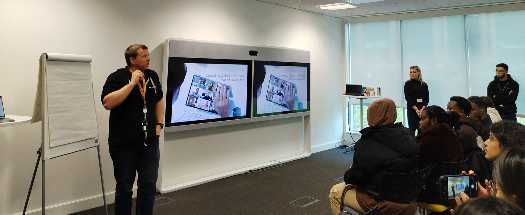 Students sit behind desks while a Cisco employee gives a presentation at the front of the room on two big digital screens.