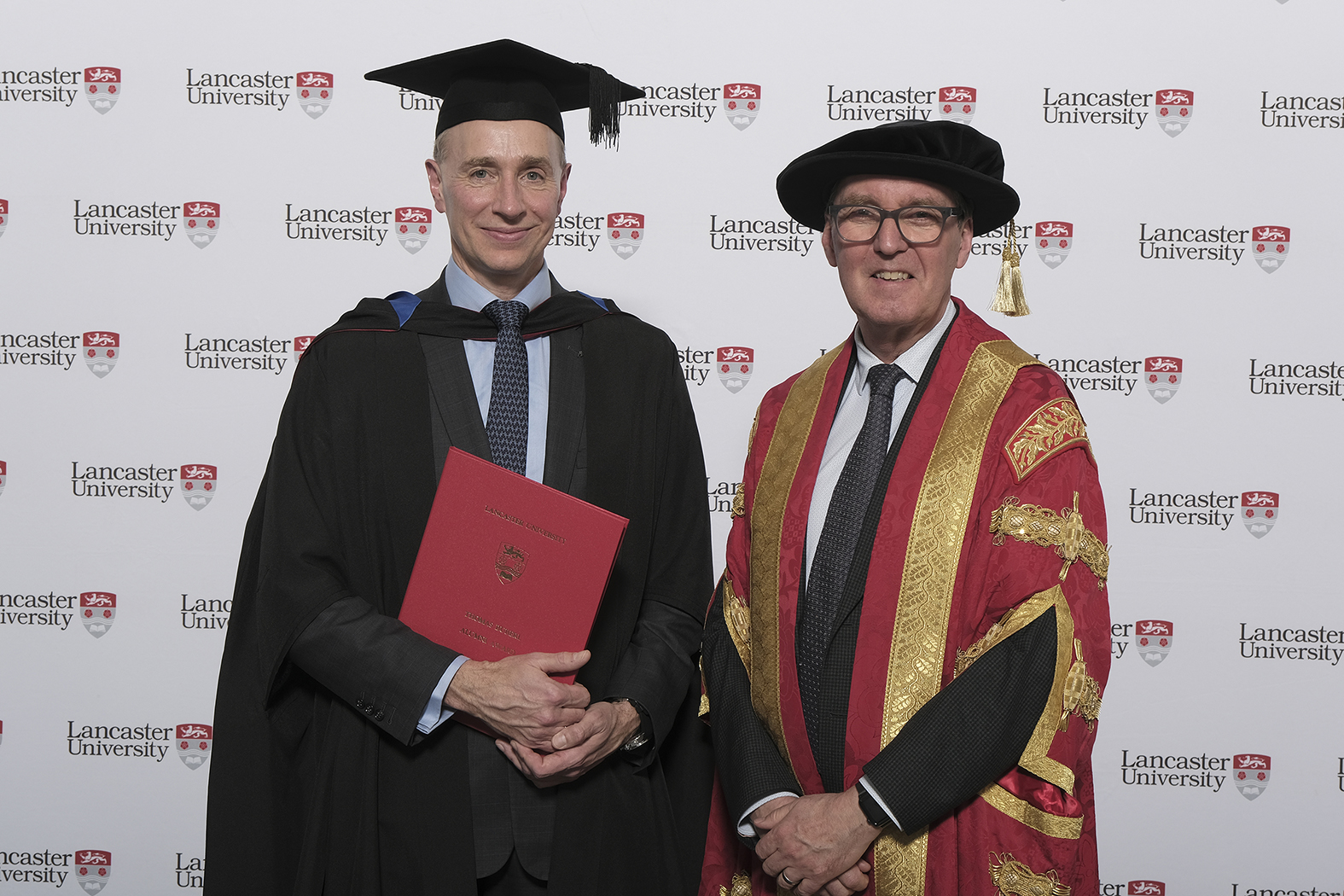 Thomas Buberl (left), in graduation gown and hat, stands with University Chancellor, the Rt Hon Alan Milburn in front of a background decorated with the Lancaster University logo.