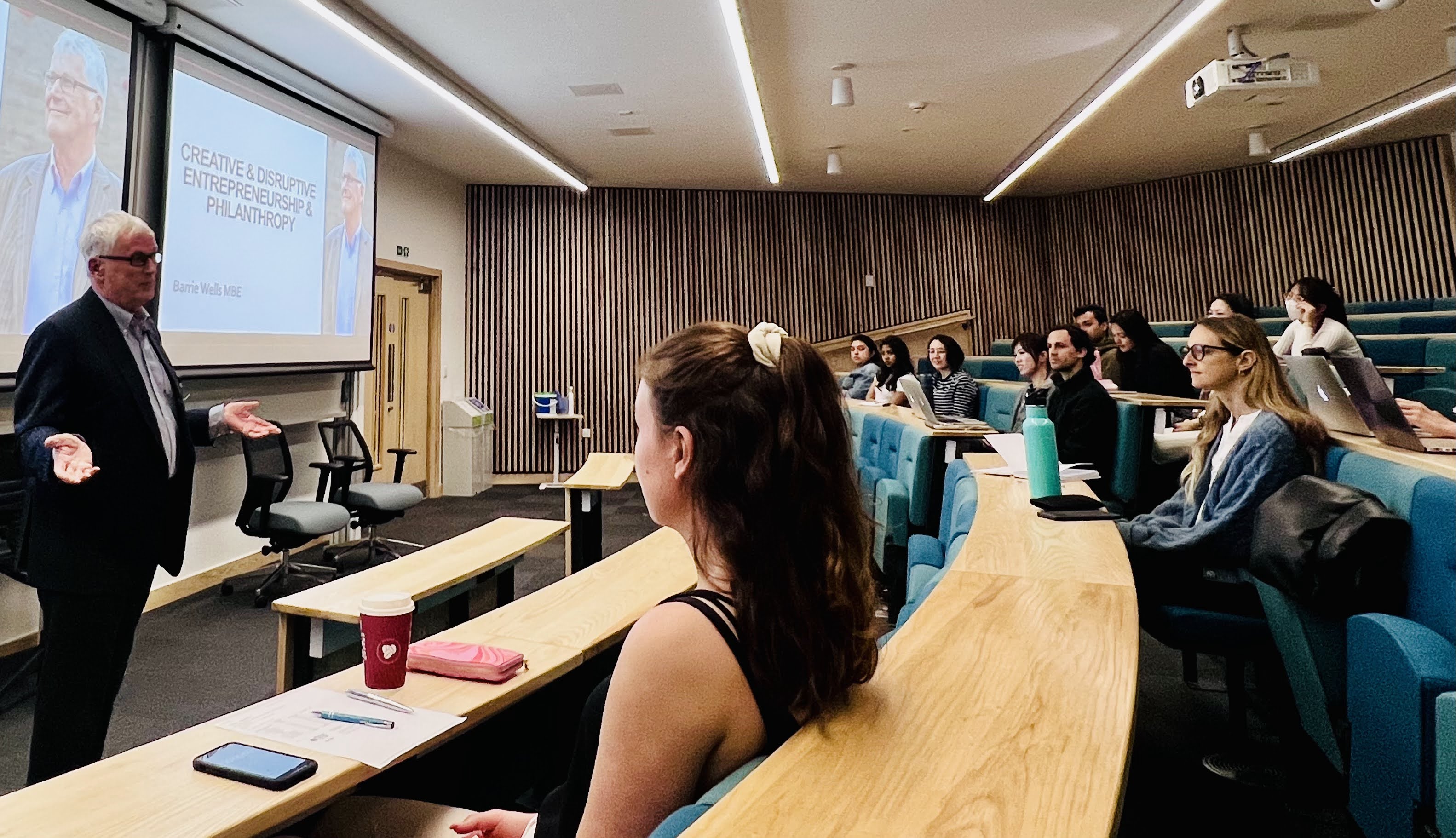 Barry Wells, Lancaster University Management School Leader in Residence, speaks to students on the MSc Digital Business, Innovation and Management programme. He is stood at the front of a leacture theatre, with students sat in curved rows of cushioned wooden seats.