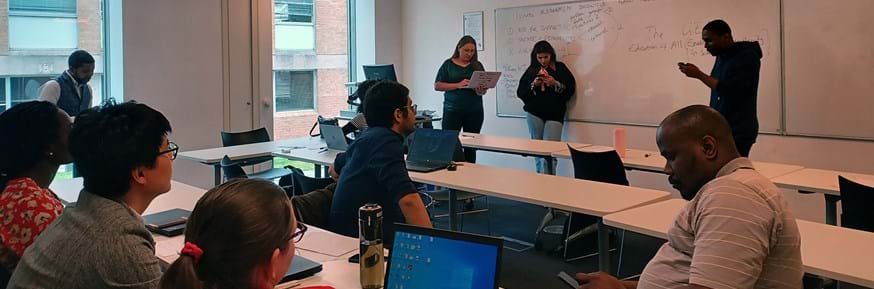 A group of early career researchers sat at desks and stood in front of a whiteboard in a seminar room as part of a research grant writing event.