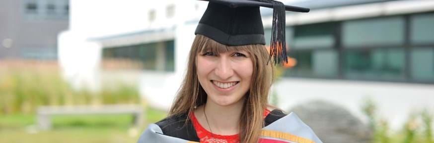 Smiling Eleanor Tinsley in graduation gown and mortarboard hat in one of the Lancaster Environment Centre garden areas