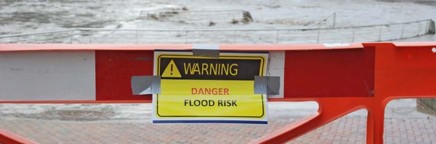 A red barrier in front of a flooded river with a bright yellow sign on it saying 