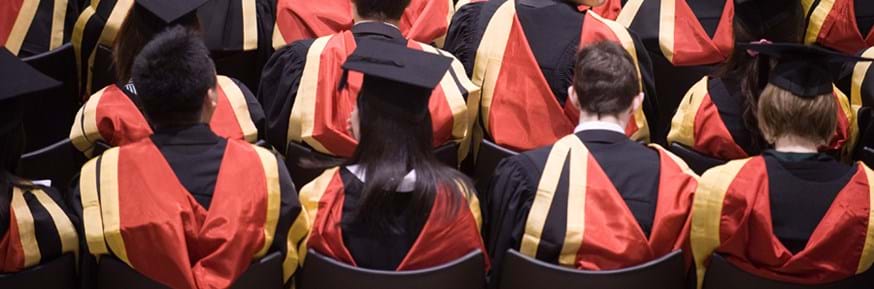 A view from above and behind a row of students in black gowns red and gold hoods down over their shoulders and mortarboard hats on their heads 