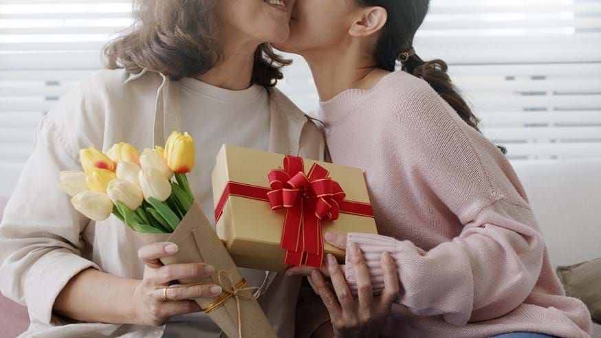 Image of an adult daughter kissing her mother and handing her a gift and some flowers