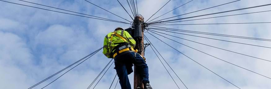 An image of a telecoms engineer wearing hi-vis working at height on a telecoms pole, set against a bright blue sky