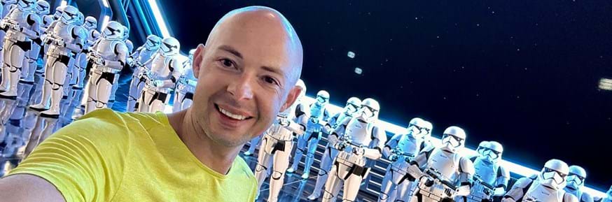 Antony Chesworth, wearing a yellow t-shirt, takes a selfie in front of a line of Star Wars Stormtroopers