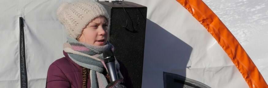 Pale skinned young girl in woollen bobble hat speaking into microphone, in front of white arctic shelter