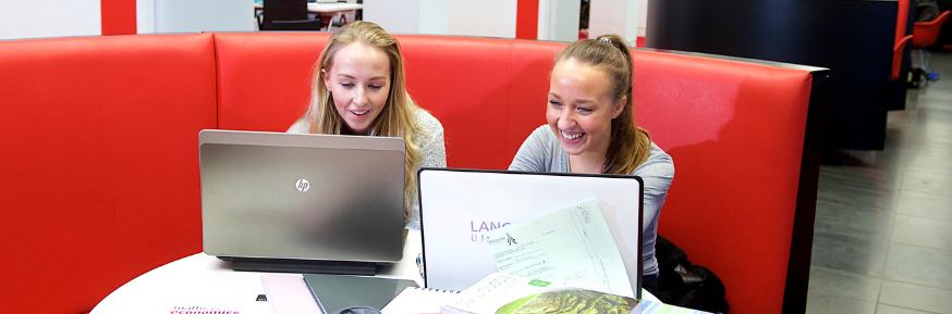 Photograph of 2 women working on their laptops and laughing in the Learning Zone on Lancaster University Campus.
