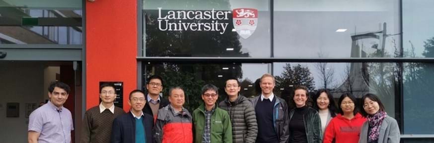 A diverse group of people standing in front of a large glass window, beneath the Lancaster University logo