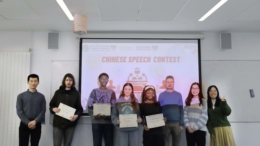 A group of people stood smiling at the camera. The four students are holding a certificate. Behind them is a slide that says 'Chinese speech contest'