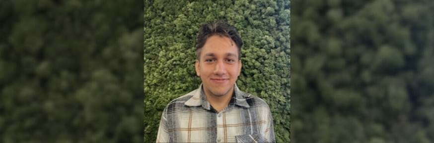 Profile photograph of Monib. He wears a check shirt and is standing in front of a green, natural-looking backdrop
