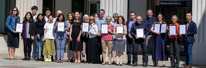 Award winners from the Lancaster University Management School Dean's Awards 2023 stand in a line holding their certificates outside Lancaster University Management School. The words 'management school' are written in large metallic writing above the building entrance.