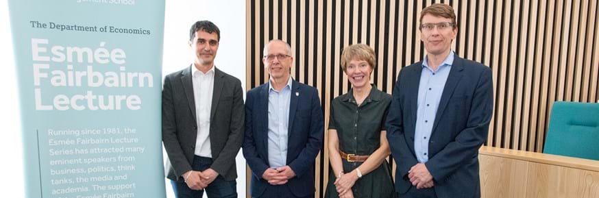 From left: Professor Themis Pavlidis, Professor Steve Bradley, Professor Claire Leitch and Professor Klaus Adam stand side-by-side in front of a wooden slatted wall and next to a light blue banner saying 'Department of Economics: Esmee Fairbairn Lecture'.