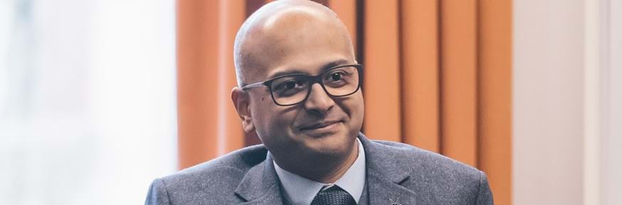Nahim Choudhury, wearing a grey suit, is sat down and smiling during one of the Help to Grow sessions, looking slightly to the right of the camera