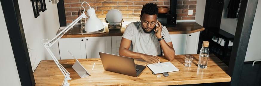 A black man with glasses and in casual dress working at a desk from home, sat in front of a laptop and holding a mobile to his ear