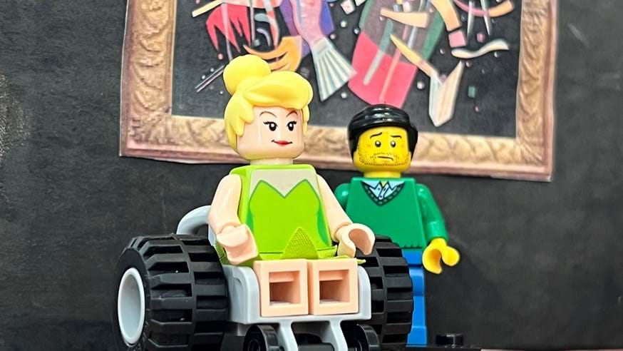 Two Lego figures, one in a wheelchair and one standing, in front of a miniature artwork in a mock-up of an art gallery
