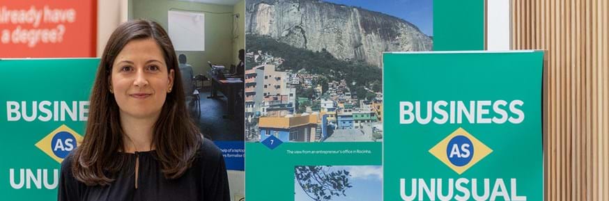 Dr Josiane Fernandes stands in front of a number of colourful panels displaying her photographs of the favelas in Rio de Janeiro. The panels are in the colours of the Brazilian flag - green blue and yellow. Text on the front panel reads 'Business as Unusual: A Photographic journey through the favelas of Rio de Janeiro by Josiane Fernandes'.