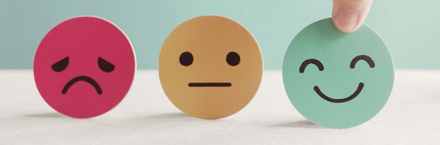 3 circle paper cut out's with happy, sad and neutral emotions depicted on them, with a hand choosing the happy smile face cut out