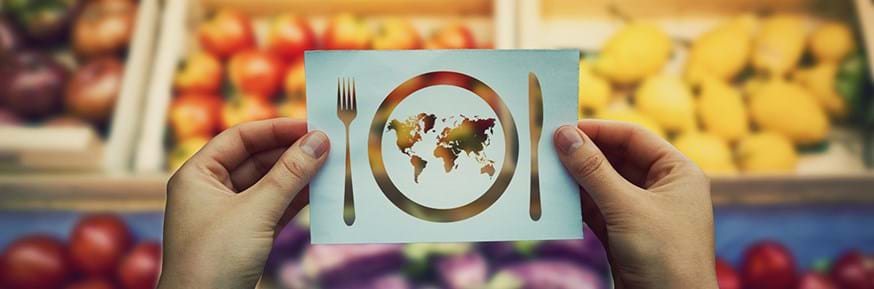 Cut-out of world map as plate with knife and fork held over market of groceries