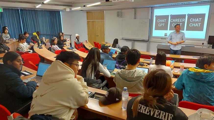 Justin Ablett stands at the front of a lecture theatre filled with students. On the screen behind him are the words 'Chat GPT' 'Carer Chat' and 'Chat Chat'