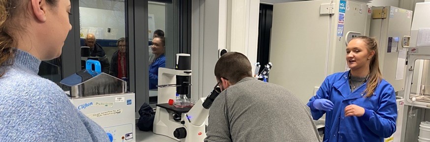 A man looks into a microscope while a researcher explains what he can see