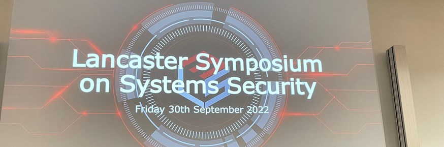 4 PhD students standing in front of a projector titled 'Lancaster Symposium on Systems Security