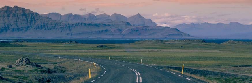 A road curves off to the left, across a landscape which is green, but very flat, and against a backdrop of mountains, at golden hour.