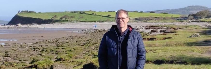 Professor Nigel Watson smiles to the camera beside a shoreline saltmarsh