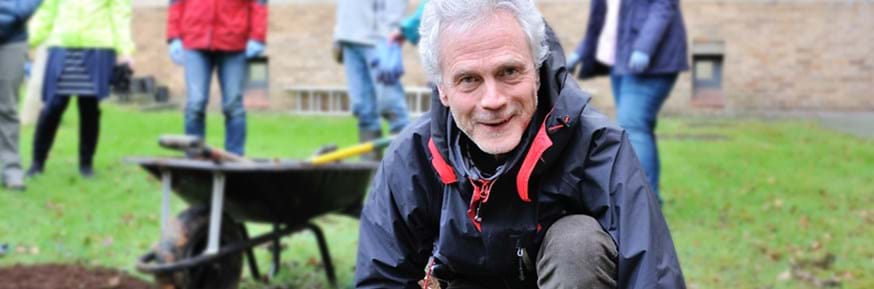 Kneeling in a lawned garden area, Nigel Paul smiles to the camera in front of colleagues and a wheelbarrow of gardening tools