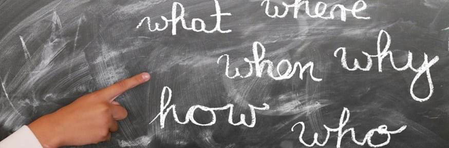 Finger pointing at a blackboard with the words what, where, when, why, how and who chalked on it.
