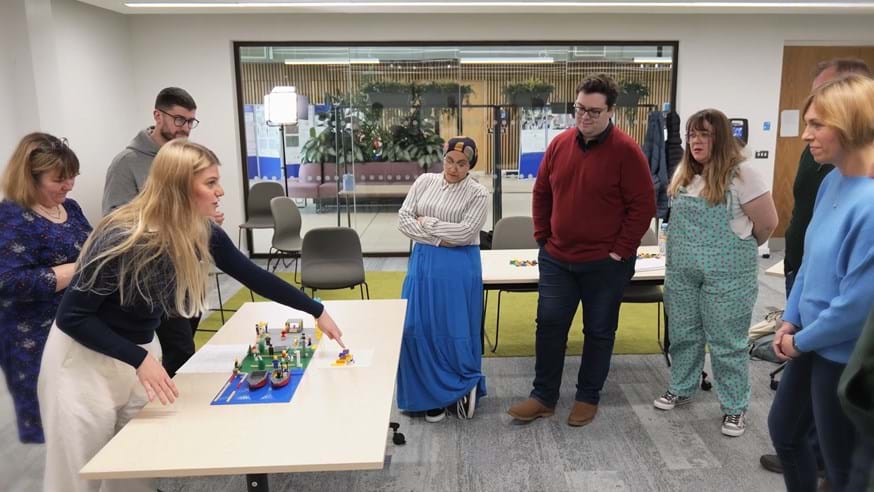 A group of people stand around a table with a Lego model on top. One of the people is pointing to a part of the model and talking to the rest of the group.