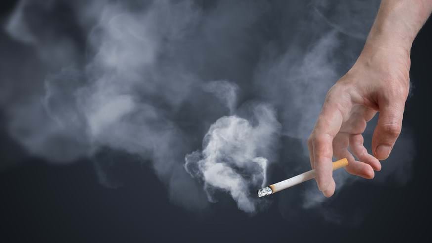 A close up image of a hand holding a lit cigarette, with white puffs of smoke set against a dark background