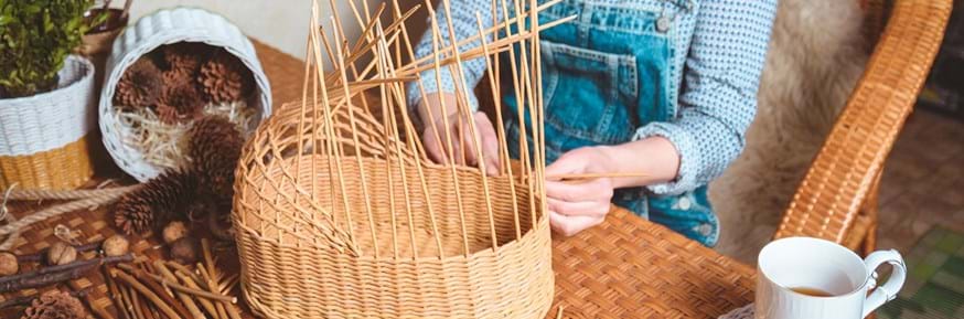 An image of a woman in dungarees sat at a table, weaving a basket