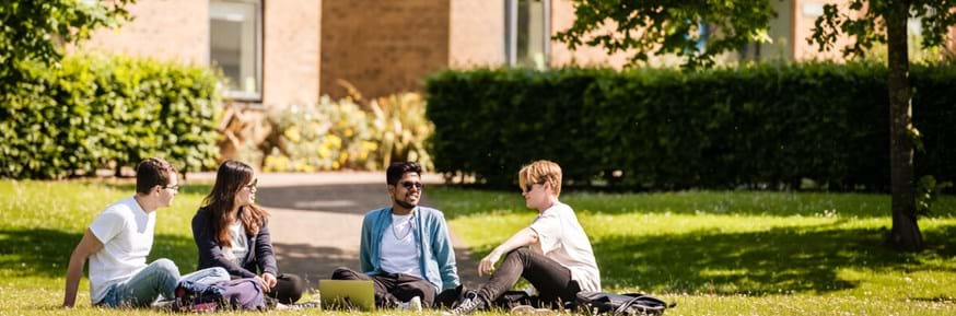Four students sitting outside on campus at Lancaster University. They are looking at each other, two are smiling. It is a sunny day and there is grass and greenery in the background.