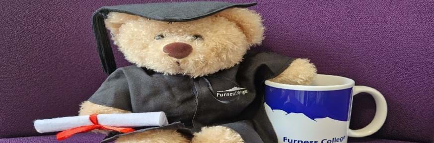 Light brown bear wearing black graduation gown, mortar hat and holding a rolled up diploma. Furness College logo printed to left side of gown.