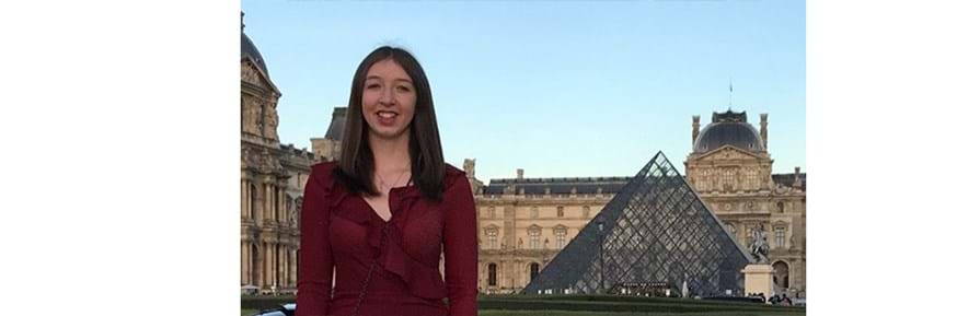 A female student wearing a magenta coloured blouse standing in front of the Louvre in Paris.