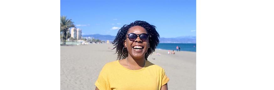 A female student standing on a beach smiling and wearing sunglasses