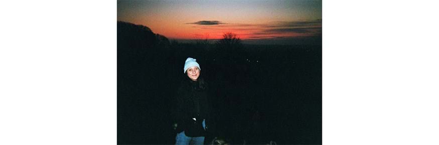 A female wearing a coat and hat in front of a sunset