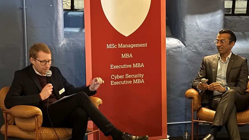 Lancaster MBA Director Dr Mark Dawson (left) holds a microphone while interviewing Vikas Shah (right). A banner in the middle lists the Lancaster MBA programmes.