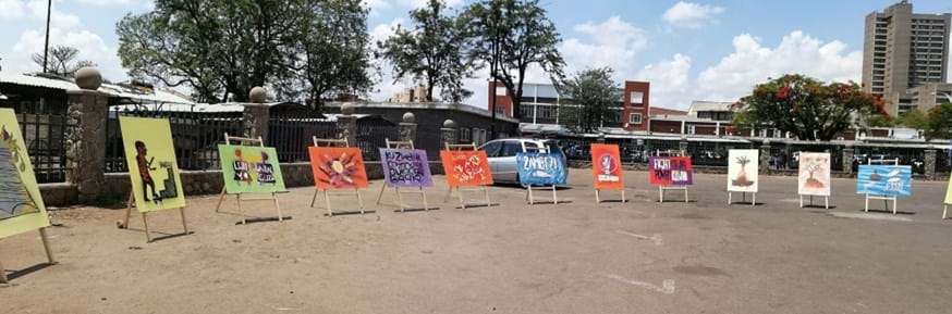 Eight easels displaying artwork produced as part of the 'Changing the story' project in Zimbabwe.  The easels are situated in an outdoor setting.